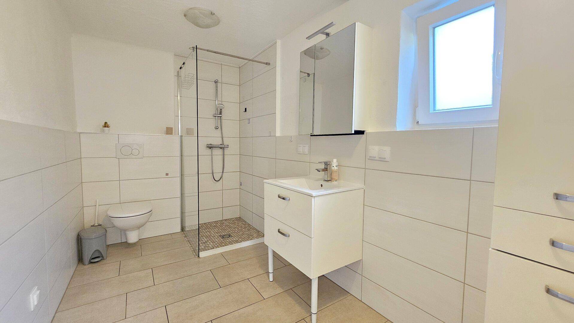Modernes Badezimmer mit begehbarer Dusche, WC und Waschtisch, hell gefliest und mit Fenster.