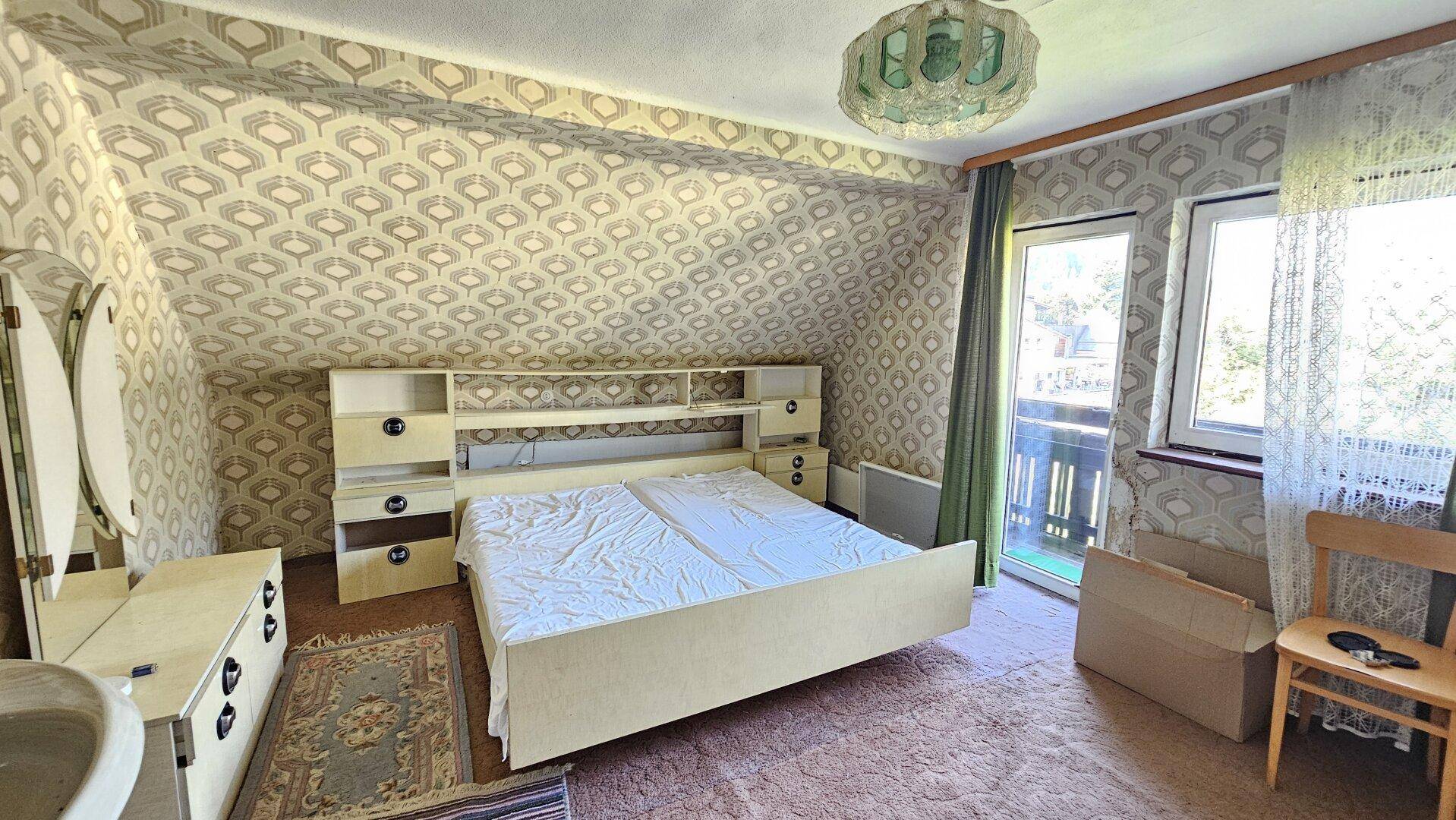Dachgeschoss-Schlafzimmer mit Doppelbett, Schminktisch und gemusterter Tapete, mit Balkonzugang.
