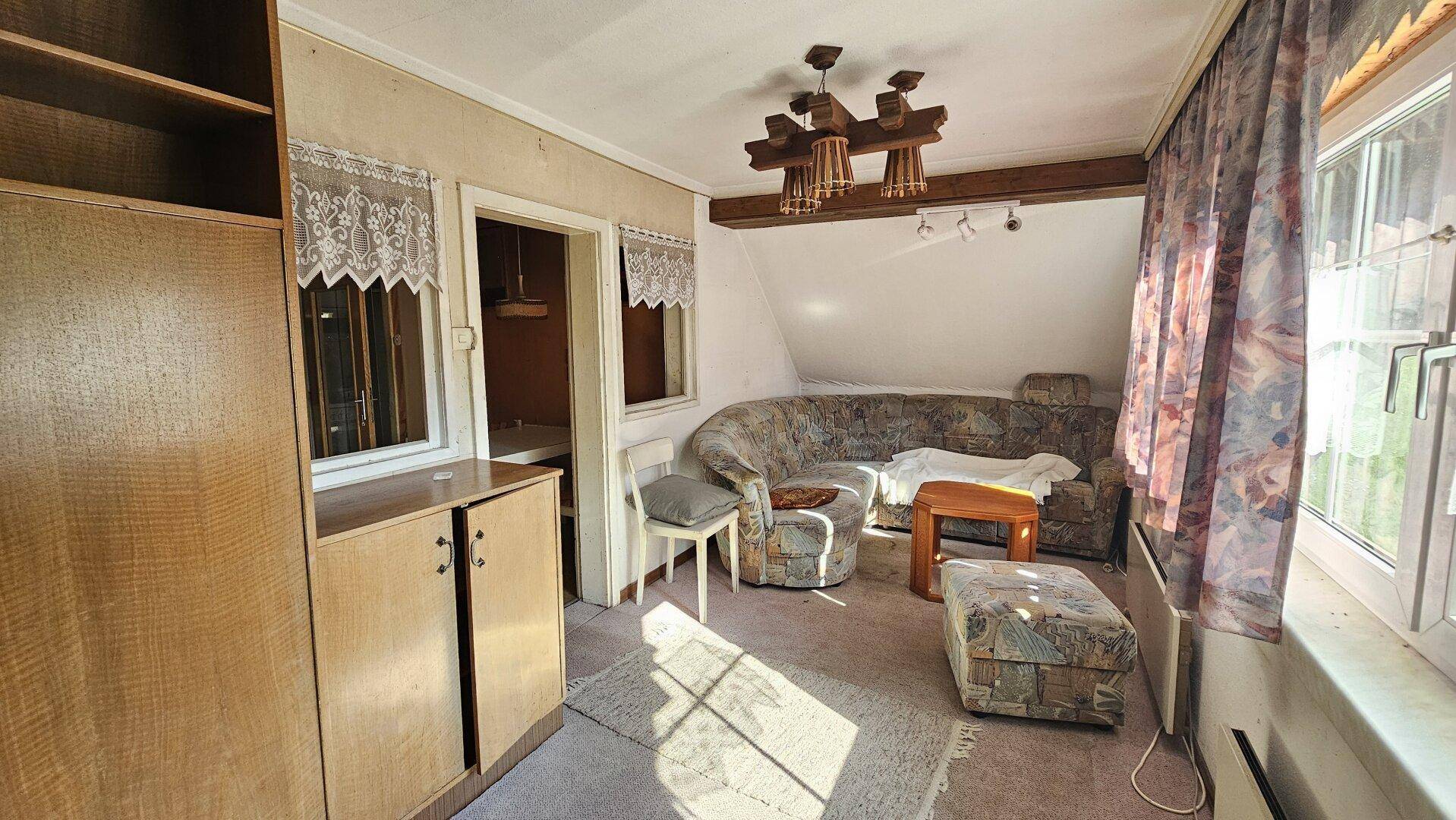 Gemütliches Wohnzimmer mit Dachschräge, Teppichboden und vintage Möbeln, durch große Fenster hell beleuchtet.