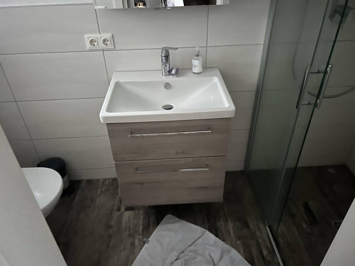 Waschbecken mit Unterschrank und Spiegel in einem gefliesten Badezimmer, neben einer Duschkabine.