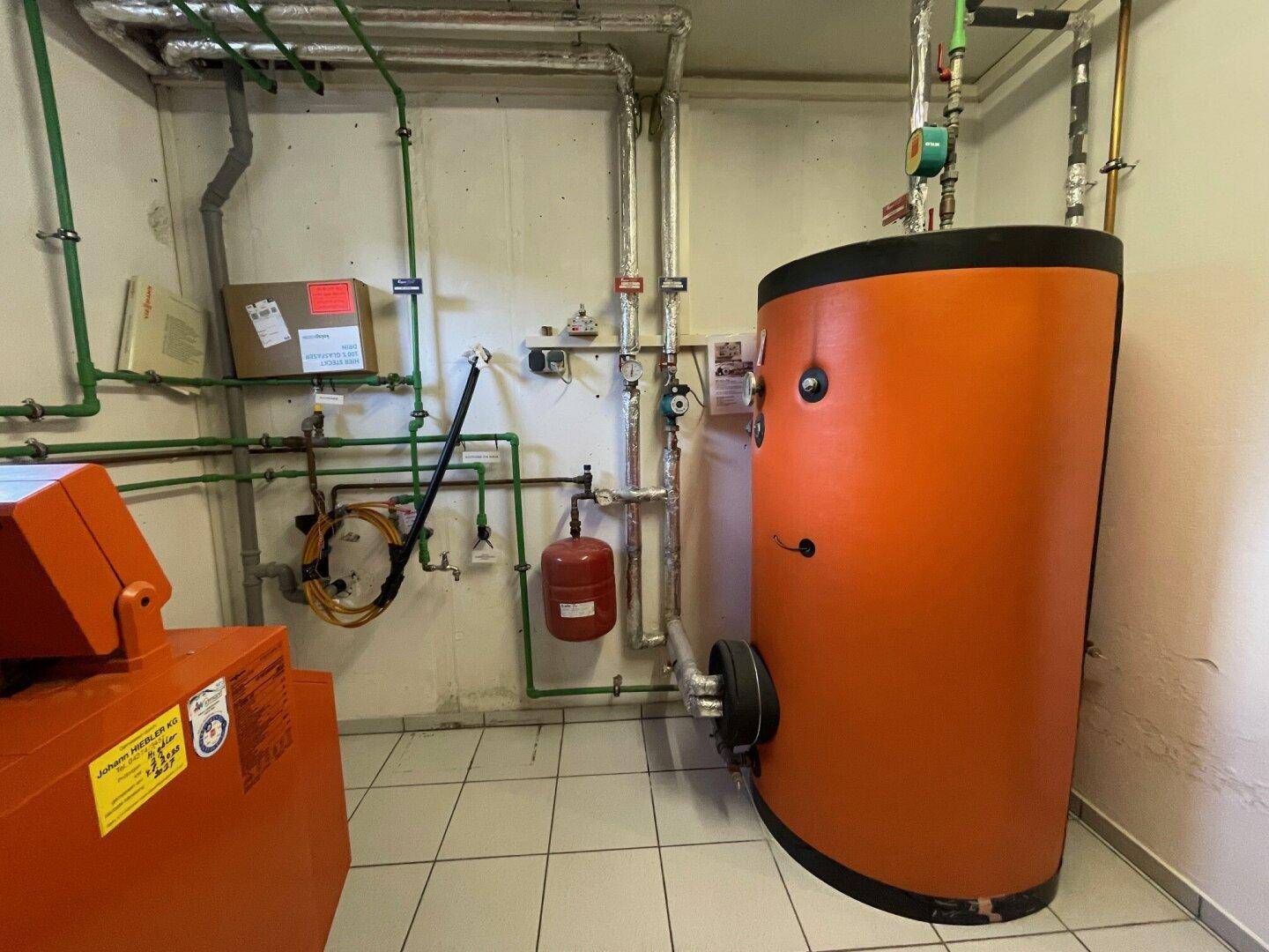 Heizungsraum mit großem Warmwasserspeicher und sichtbaren Rohrleitungen an der Wand.