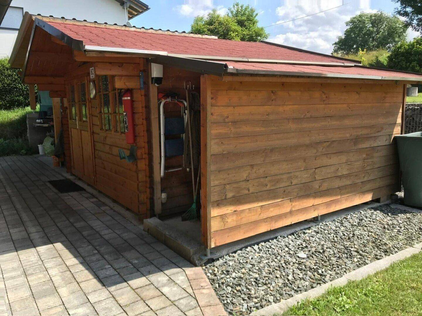 Rustikales Gartenhaus aus Holz mit rotem Dach und gepflastertem Zugangsbereich im Garten.