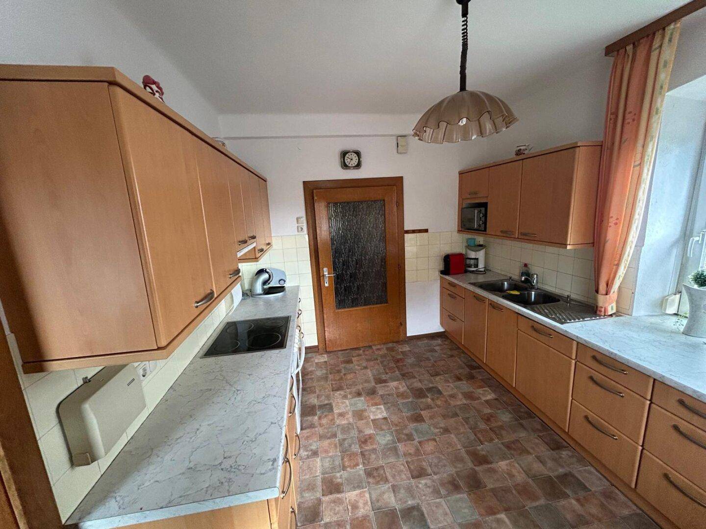 Küche mit Holzschränken, Fliesenboden und Fenster mit Vorhängen, ausgestattet mit grundlegenden Geräten.