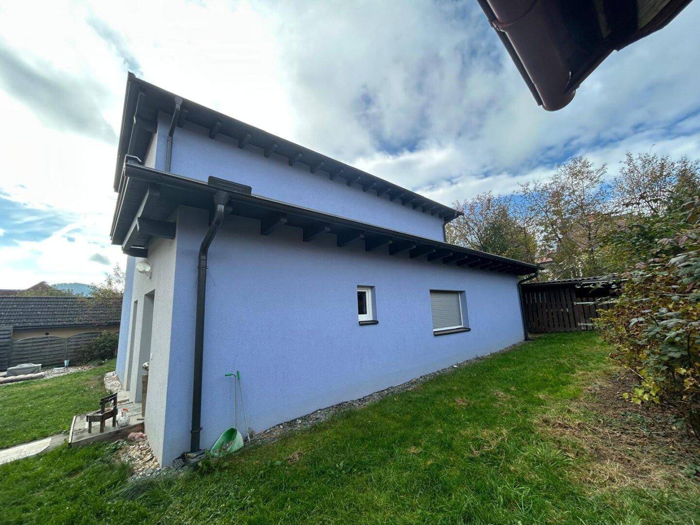Seitenansicht des Hauses mit blauer Fassade und einem abfallenden, grünen Gartenbereich.