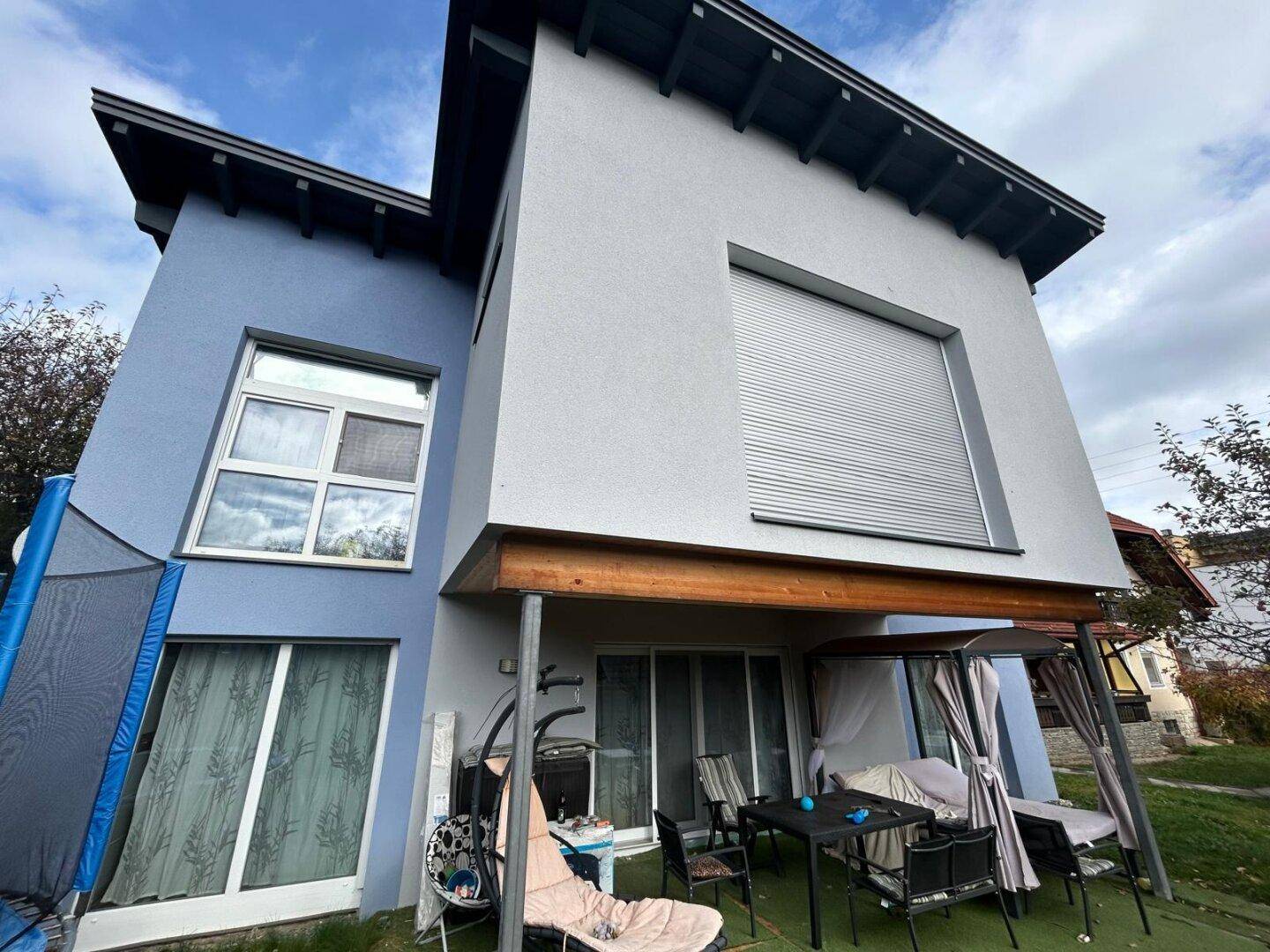 Modernes Haus mit blauer und grauer Fassade, einer möblierten Terrasse und einem Trampolin im Garten.
