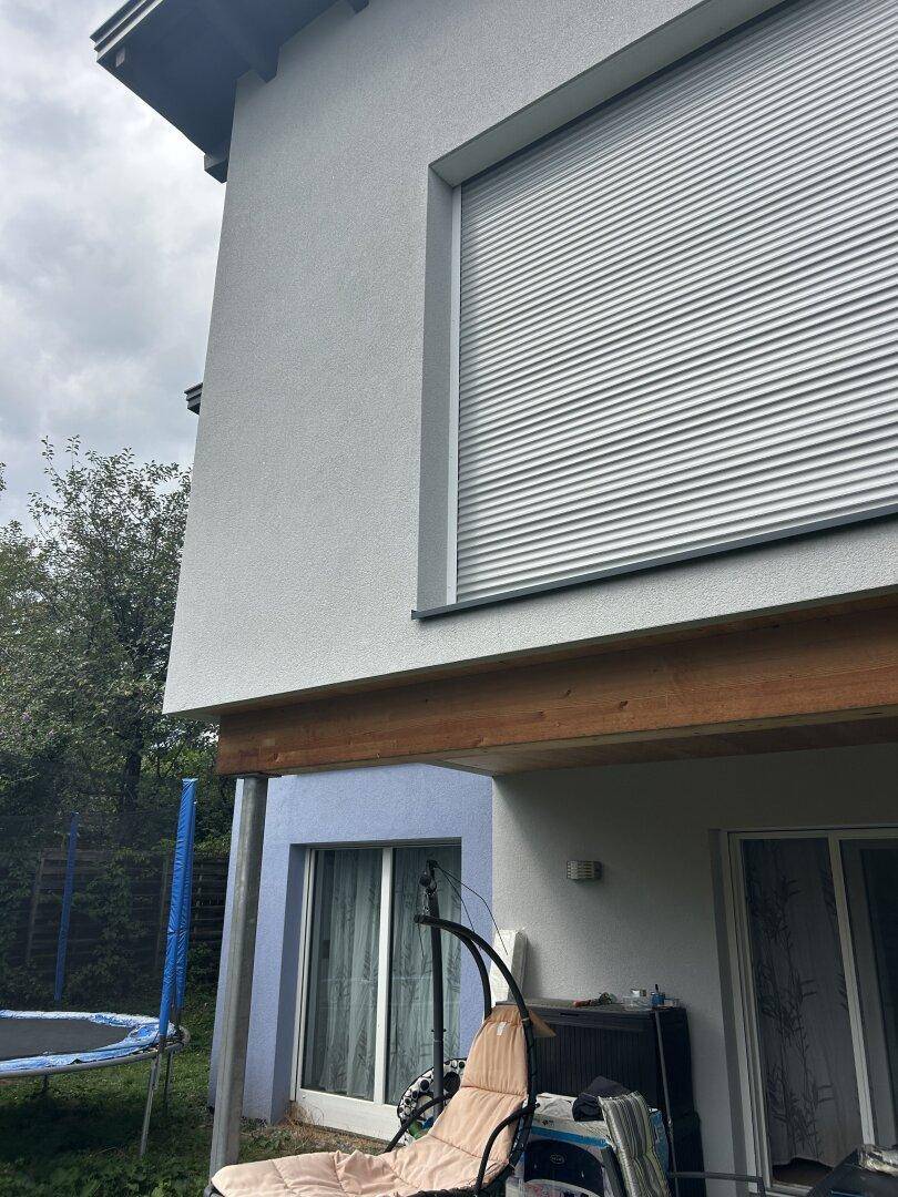 Detailansicht der grauen Hausfassade mit großem Fenster und Rollladen über der Terrasse.