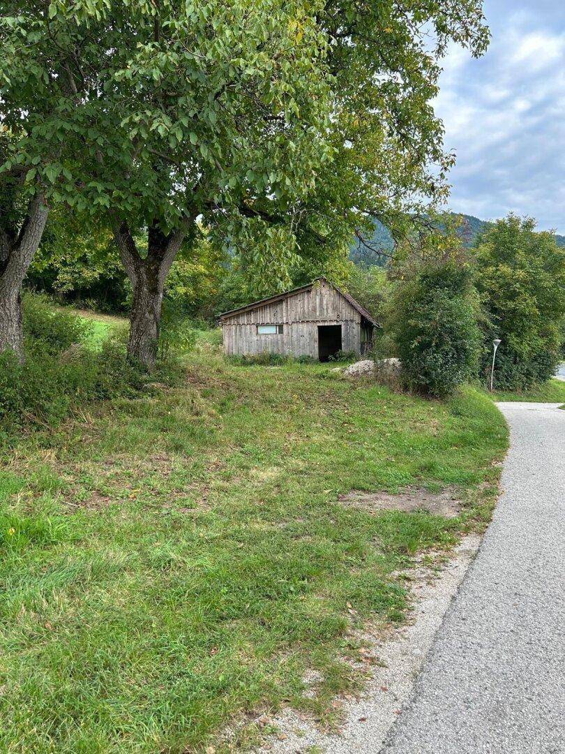 Alte Holzscheune am Rande eines ländlichen Weges, umgeben von Bäumen und Wiesen.