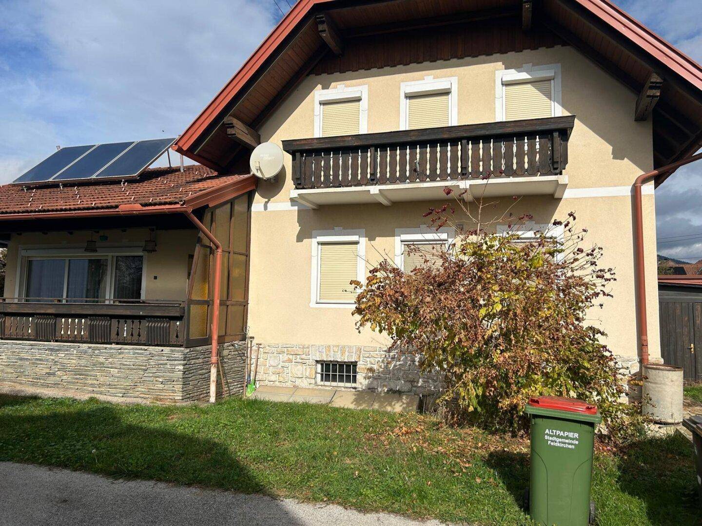 Vorderansicht des traditionellen Hauses mit Balkon, Solarpaneelen und Altpapiercontainer.