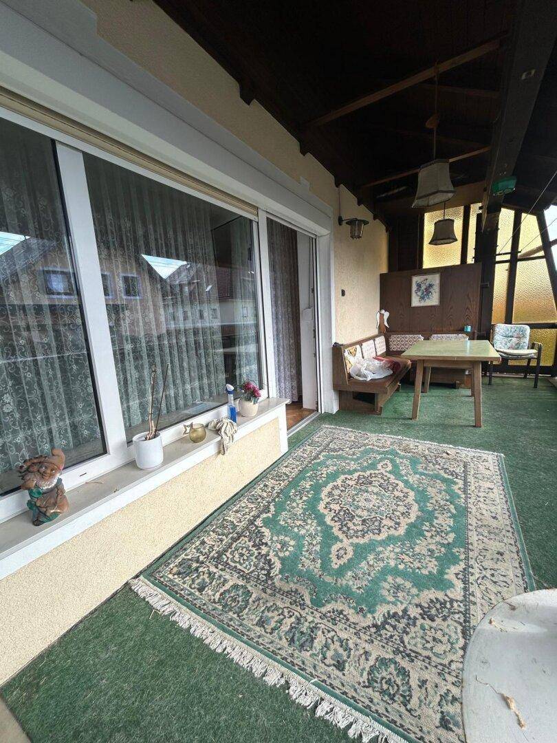 Überdachter Balkon mit Teppich, Sitzgelegenheiten und Blick ins Grüne.
