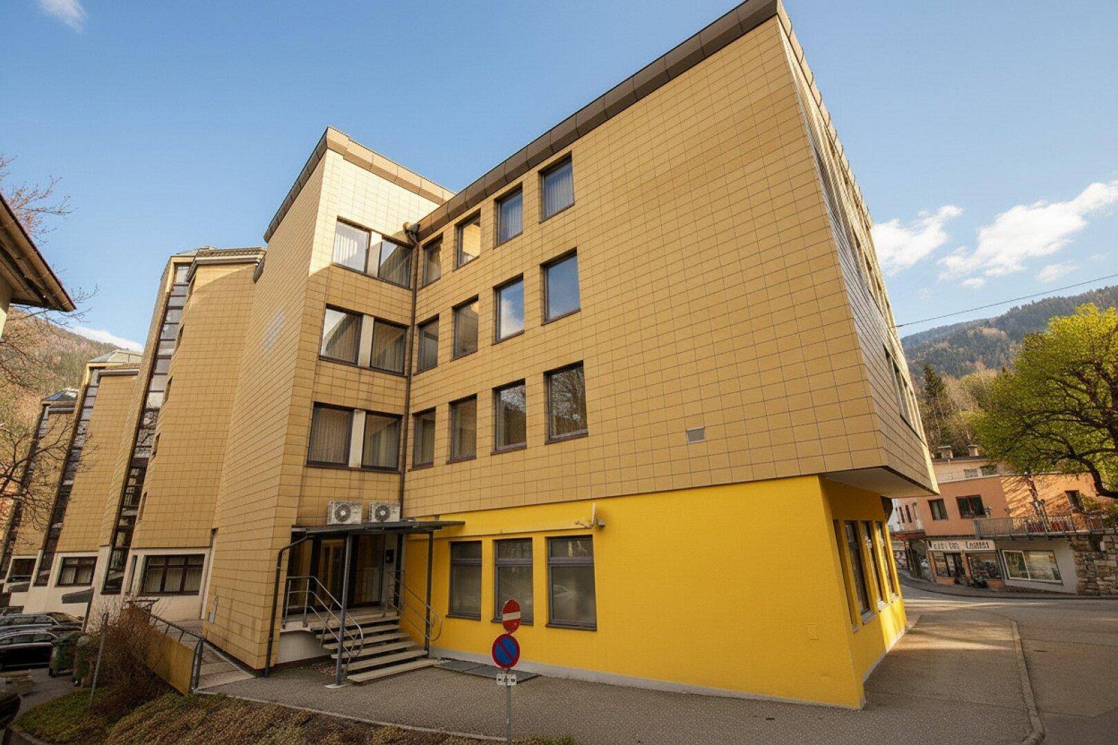 Mehrstöckiges Gebäude mit gelber Fassade und vielen Fenstern, umgeben von grünen Hügeln.