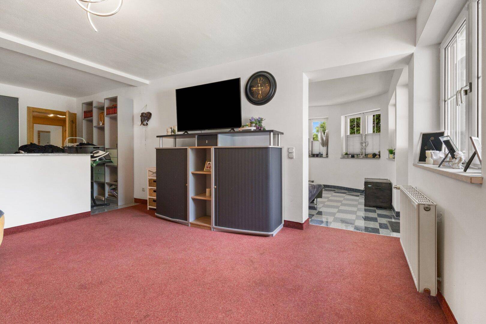 Helles Wohnzimmer mit Teppichboden, Fernseher und Blick in einen weiteren Raum.