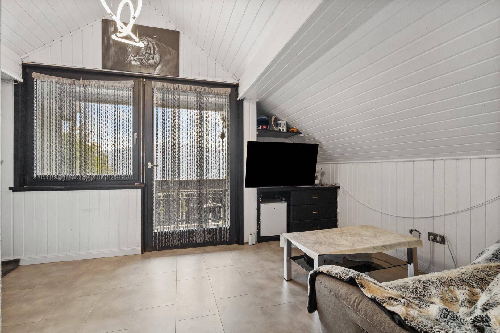 Gemütliches Wohnzimmer mit Dachschrägen, Fliesenboden und Zugang zu einem Balkon.