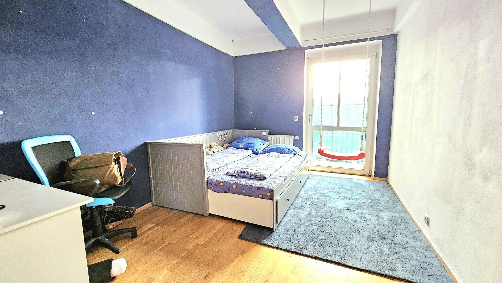 Kinderzimmer mit blauer Wand, Einzelbett und einem Schaukelstuhl am Fenster.