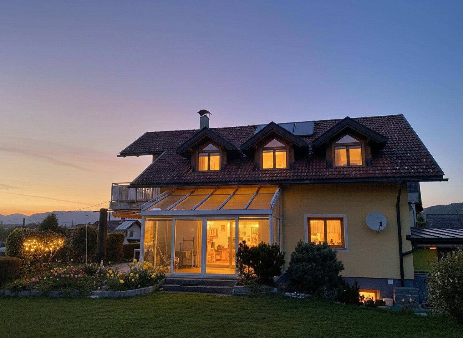 Hausfassade bei Sonnenuntergang mit beleuchtetem Wintergarten und Gartenbereich.