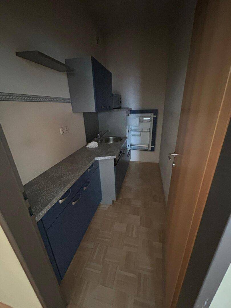 Kleine, funktionale Küchenzeile mit Spüle, Kühlschrank und Stauraum in dunklen Farbtönen.