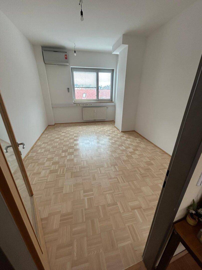 Heller Raum mit Parkettboden, einem großen Fenster und Klimaanlage, ideal für Büro oder Wohnen.