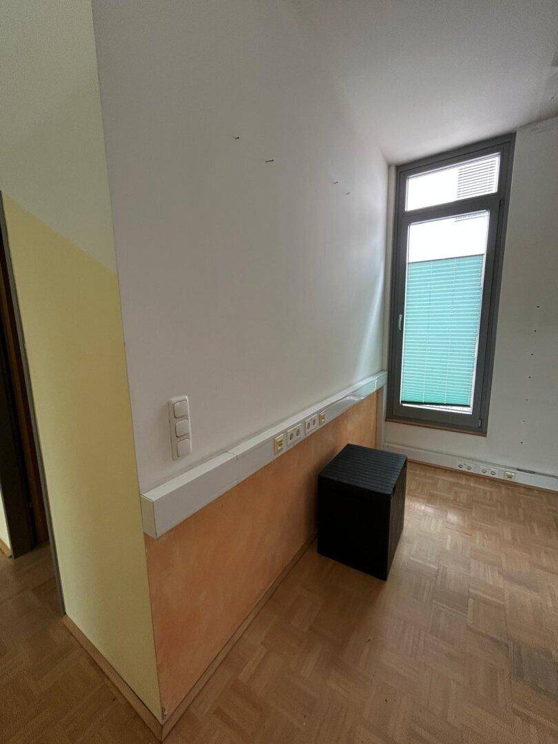 Schmaler Raum mit Parkettboden, einem Fenster mit Jalousie und Wandinstallationen.