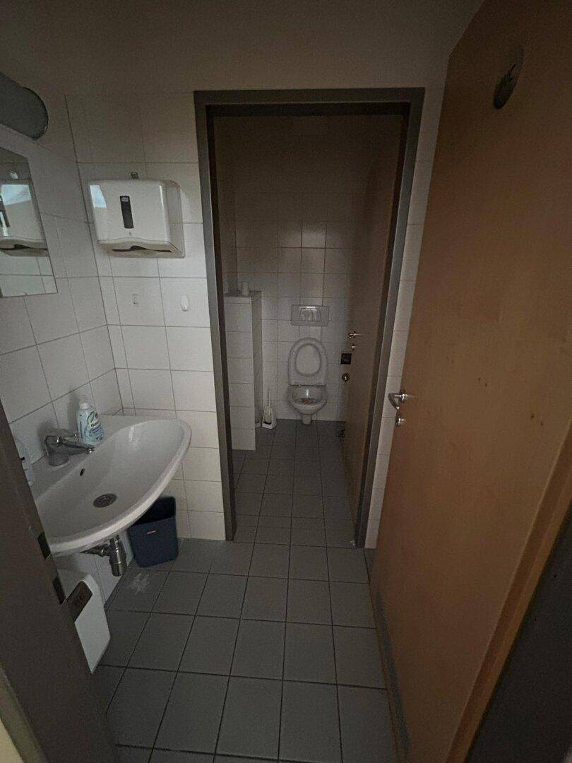 Einfache Toilette mit Waschbecken und Fliesenboden, angrenzend an einen weiteren Toilettenraum.