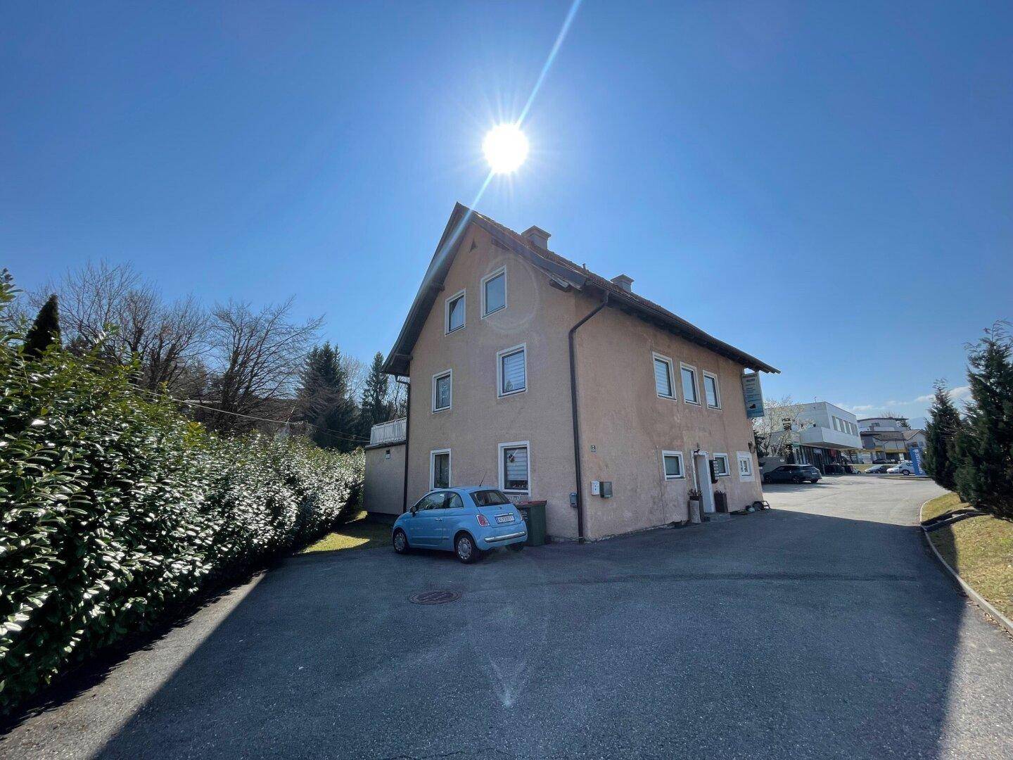 Seitenansicht des Gebäudes mit gepflastertem Vorplatz und umgebender Vegetation bei Sonnenschein.