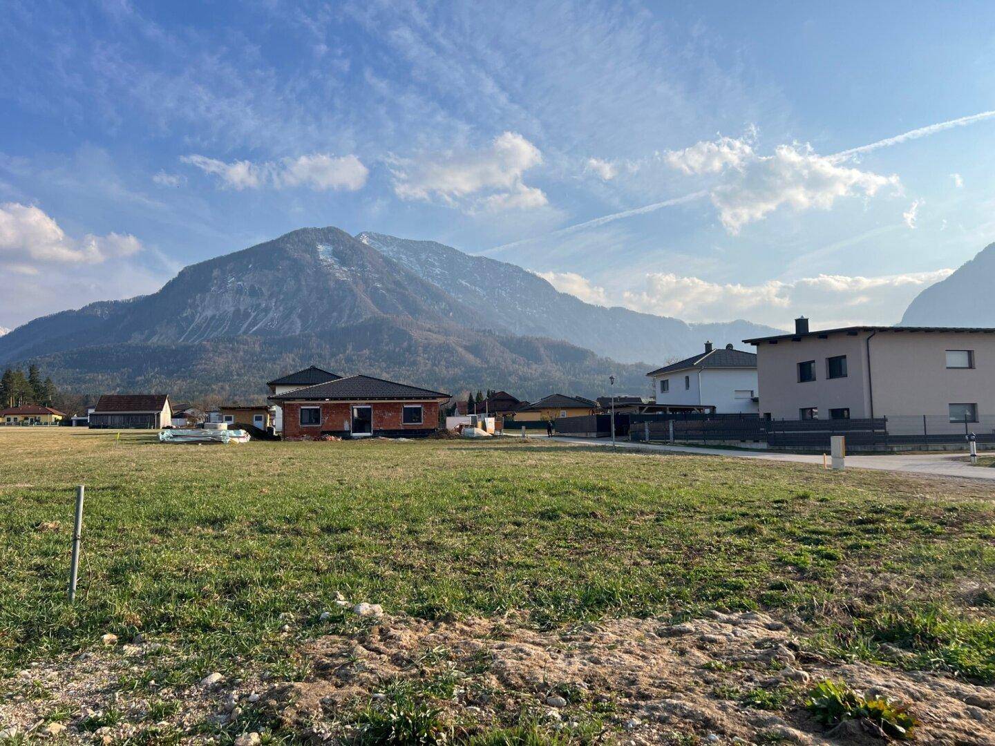 Baustelle eines Einfamilienhauses auf einem Grundstück mit weiteren Häusern und majestätischem Bergblick.