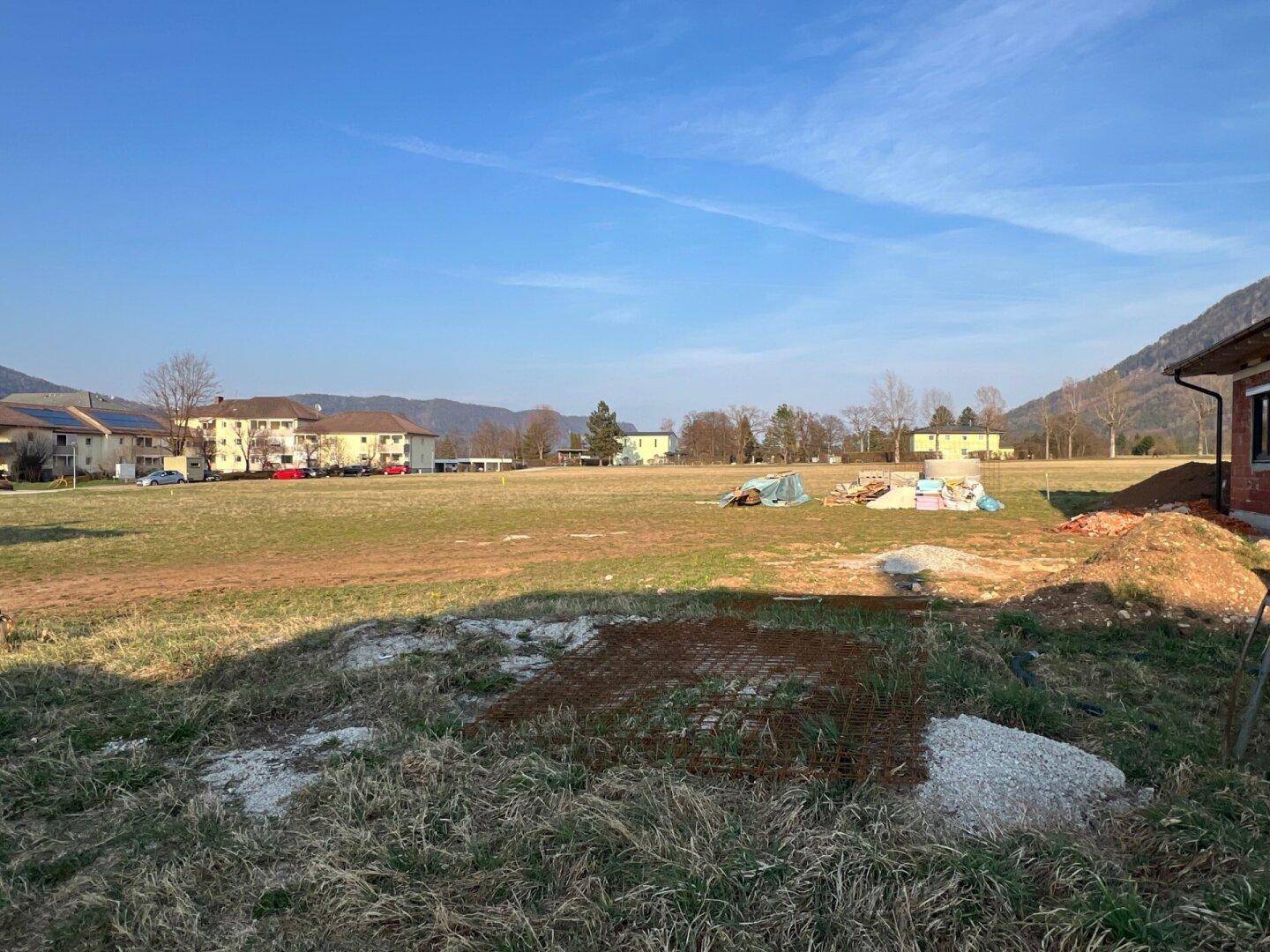 Weitläufiges unbebautes Gelände mit Baumaterialien und bestehenden Wohngebäuden im Hintergrund.