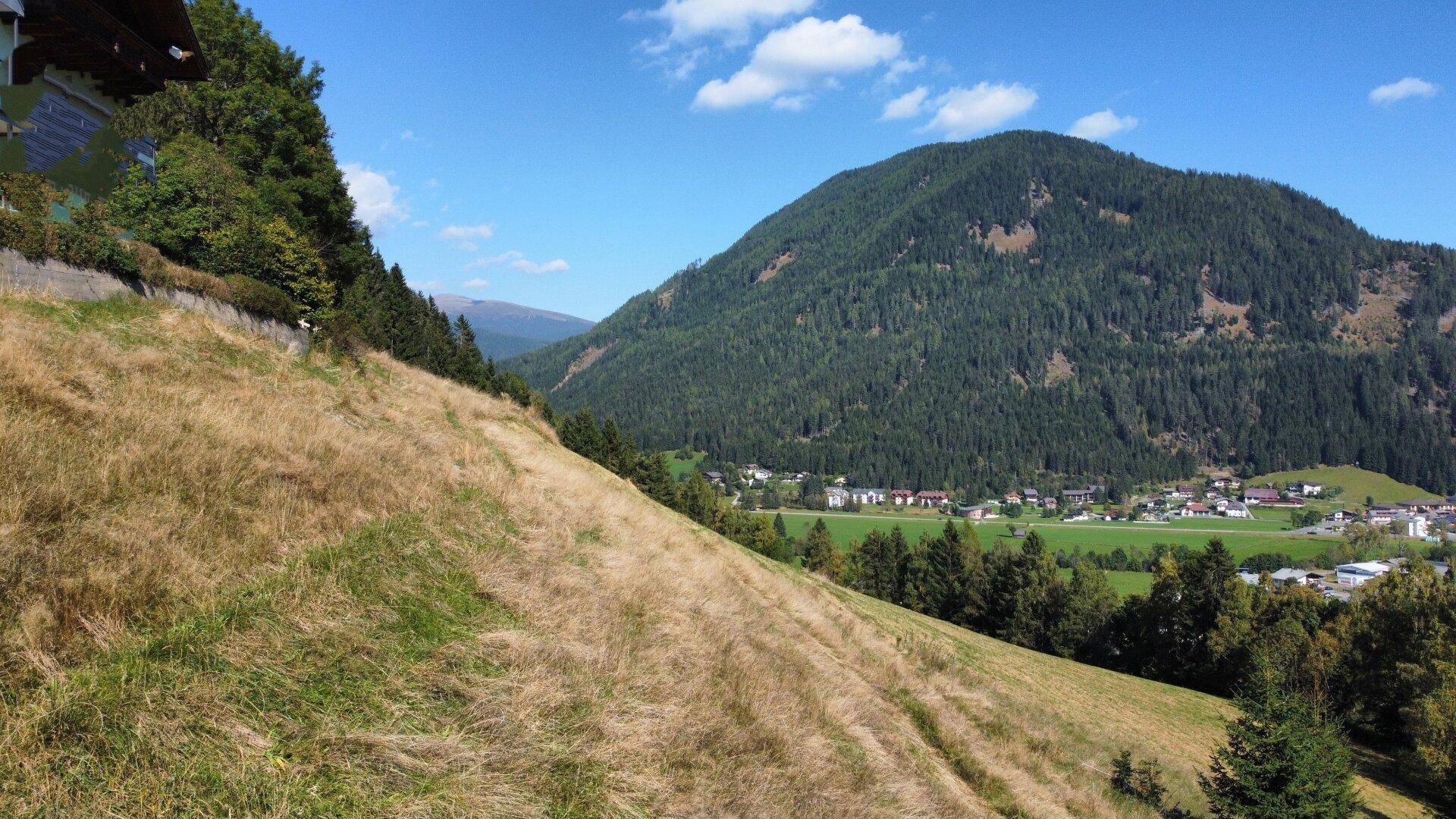 Weitläufige grüne Hügellandschaft mit Blick auf ein Tal und ein Dorf, umgeben von bewaldeten Bergen.