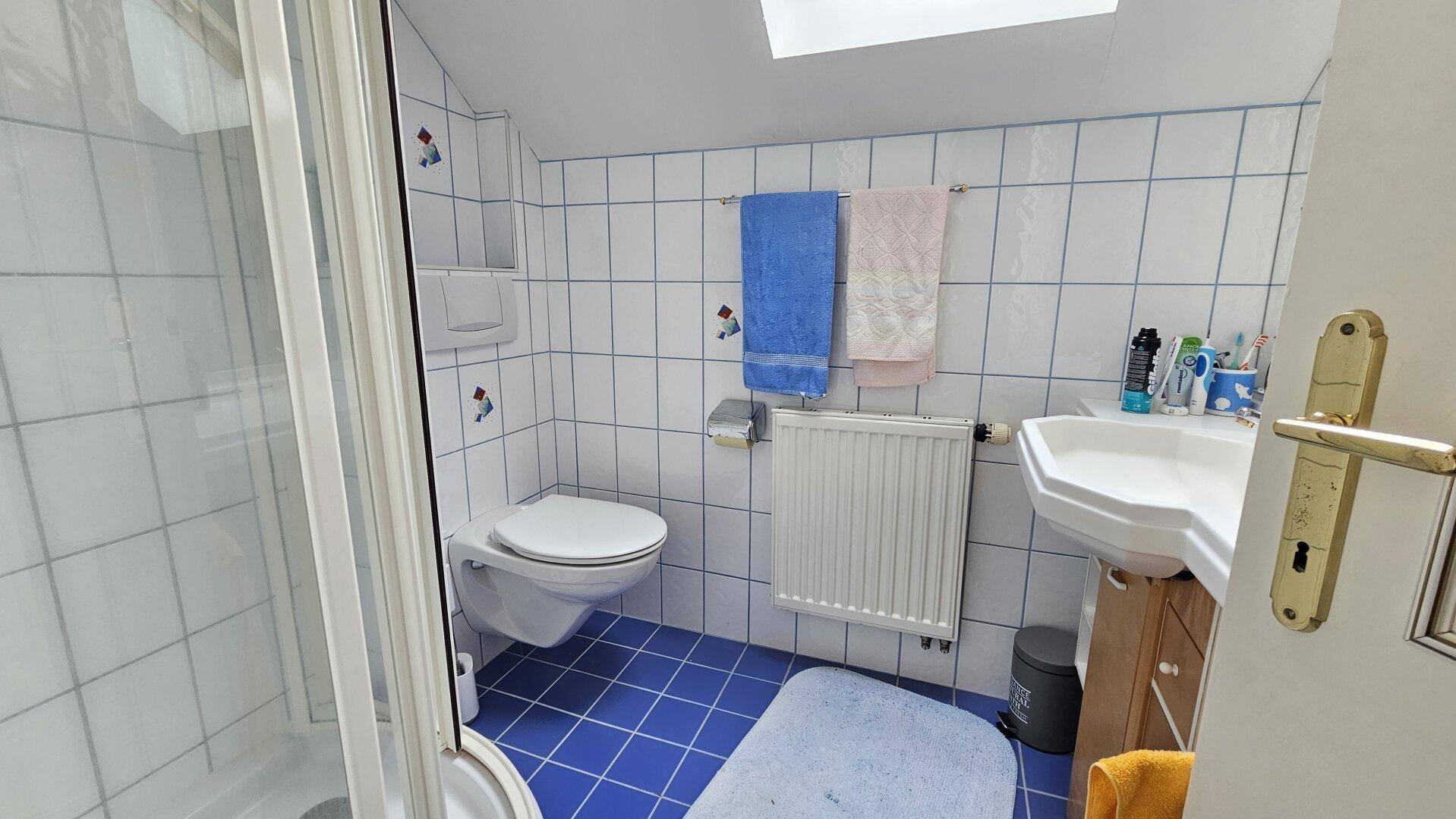 Badezimmer im Dachgeschoss mit blauen Fliesen, Dusche und Dachfenster für Tageslicht.