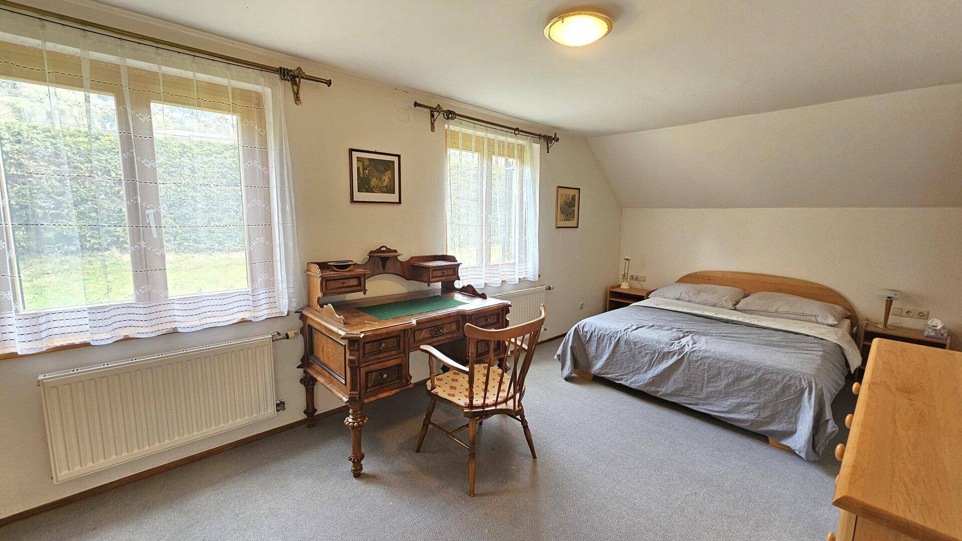 Geräumiges Schlafzimmer mit Holzboden, Schreibtisch und großen Fenstern mit Blick ins Grüne.