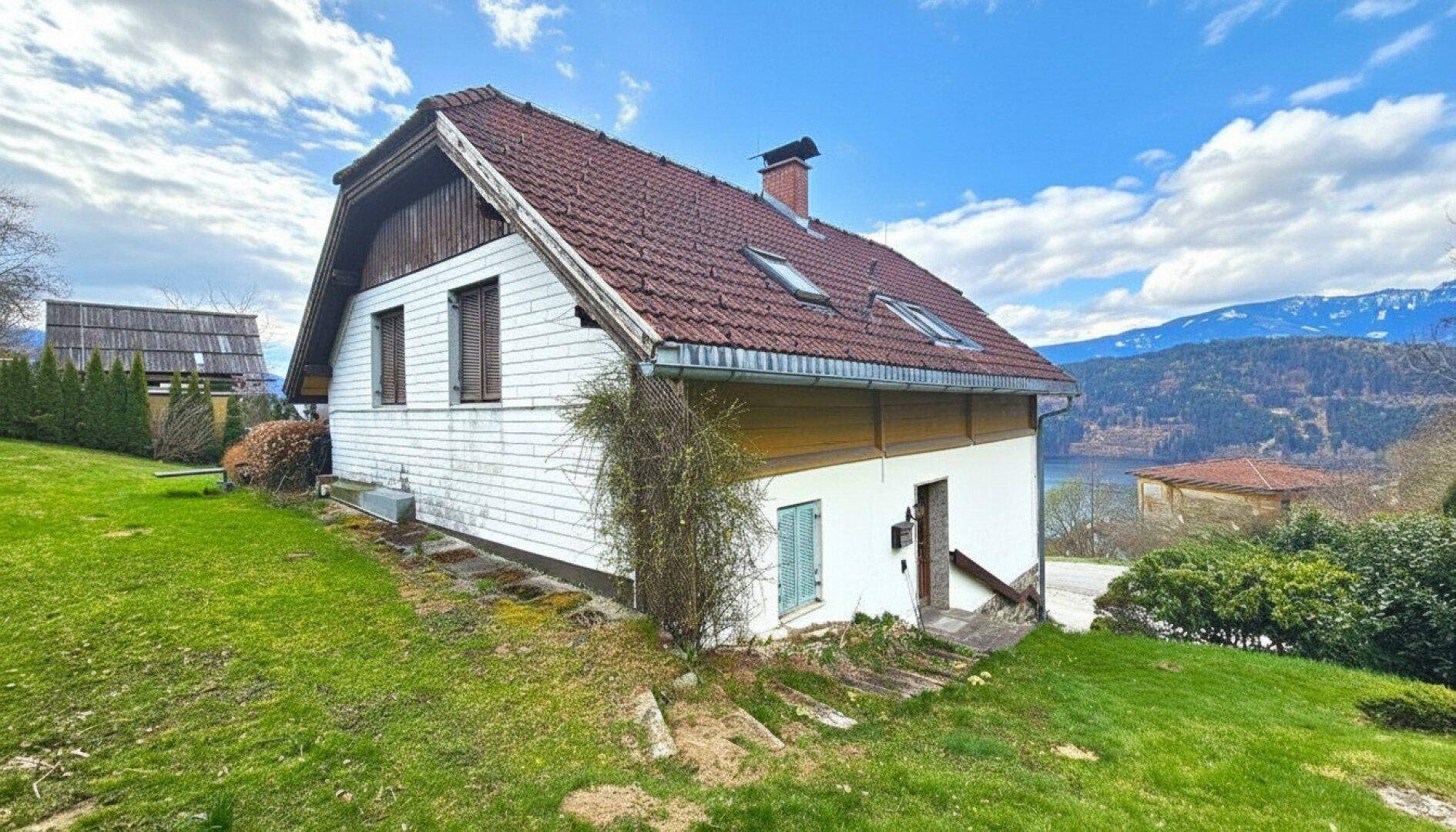 Seitenansicht des Hauses mit weißer Holzfassade, rotem Ziegeldach und Blick auf den See.