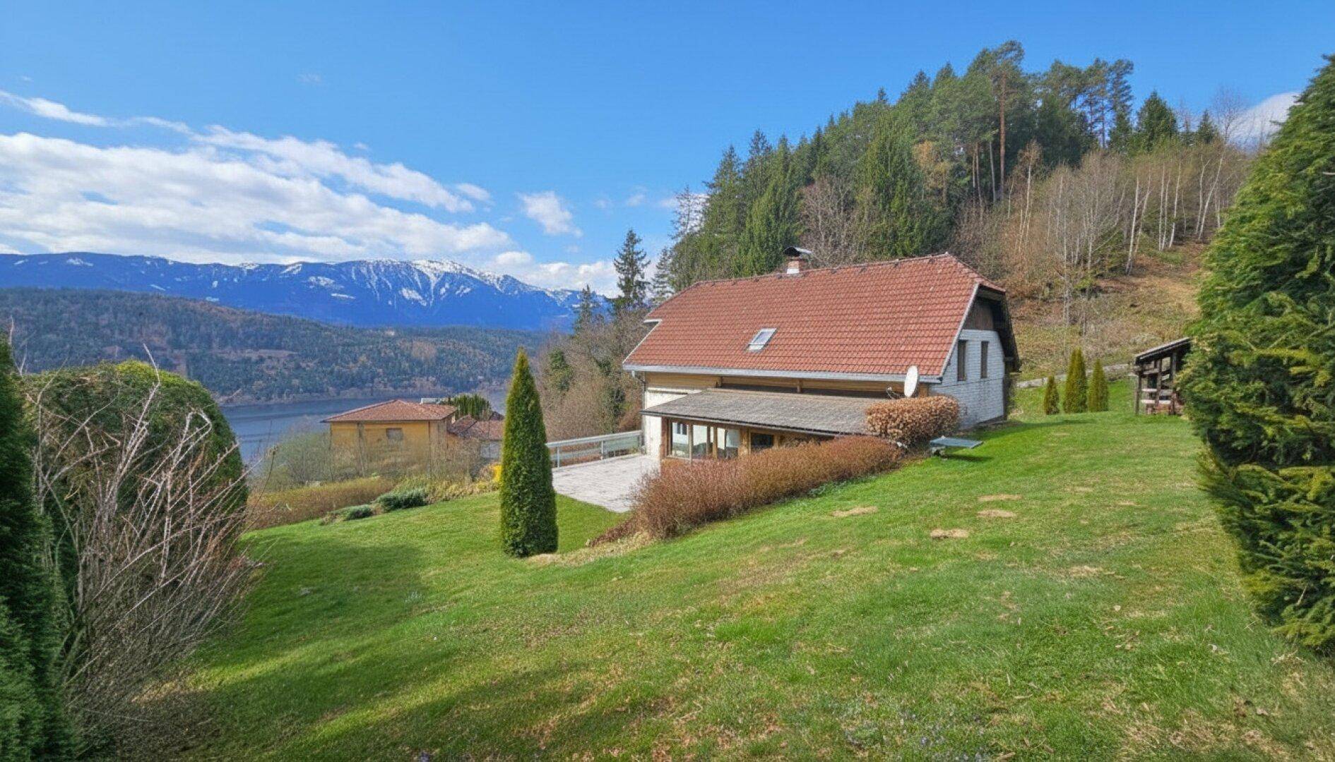 Haus mit rotem Ziegeldach und großem Garten, umgeben von Wald und mit Blick auf den See.