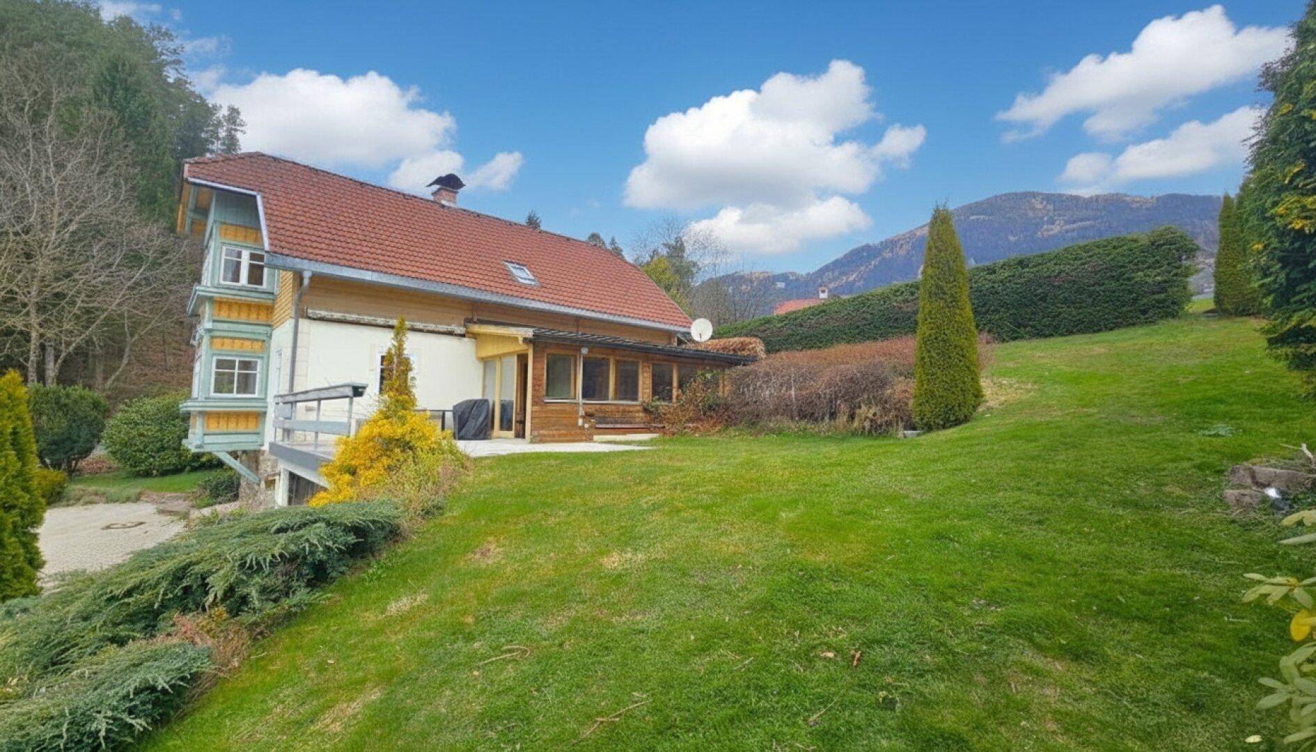 Rückansicht des Hauses mit Terrasse und weitläufigem Rasenbereich, umgeben von Bäumen und Sträuchern.