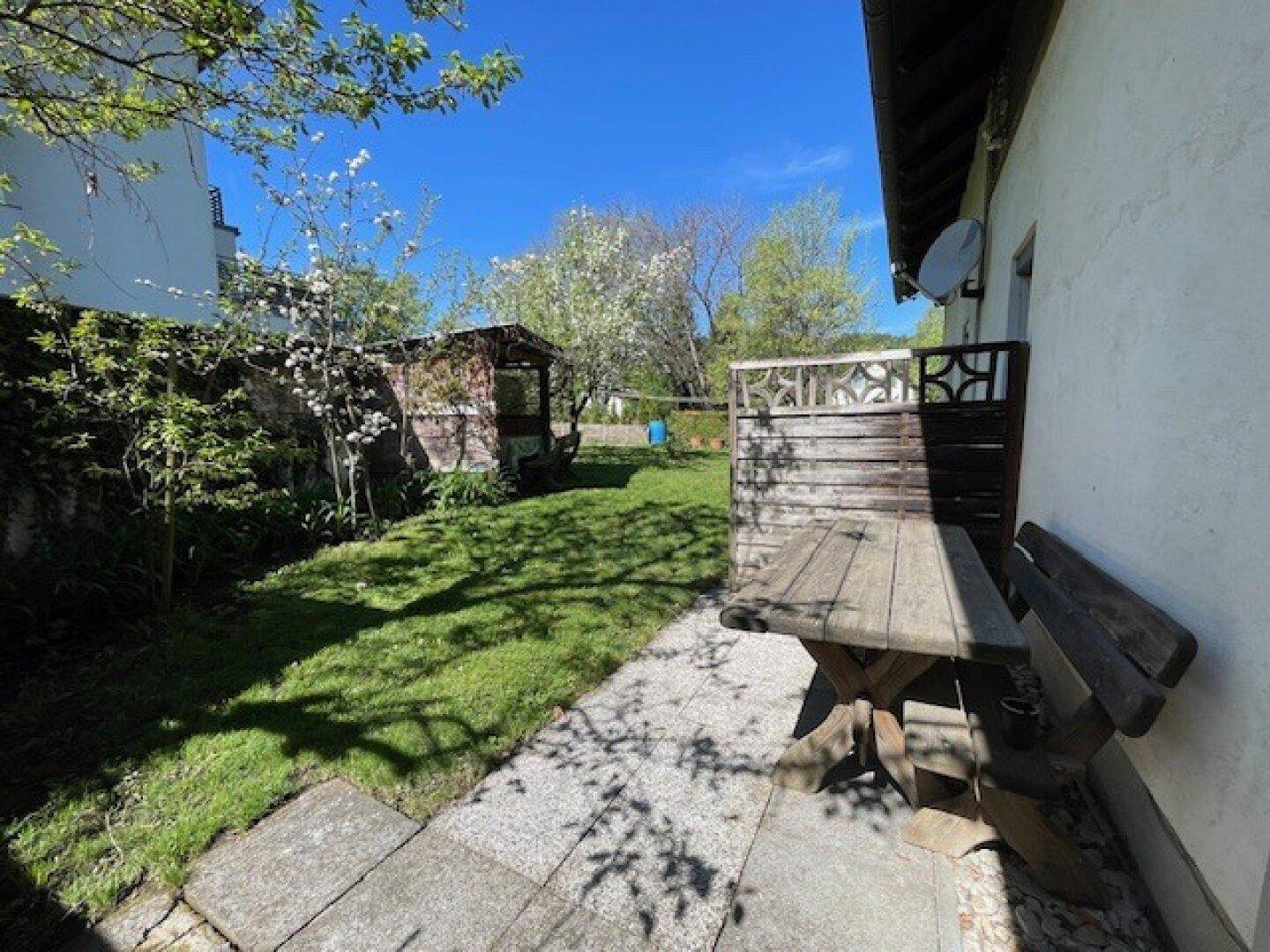 Gemütlicher Gartenbereich mit Holztisch und Bank, umgeben von Grün und einem kleinen Gartenhaus.