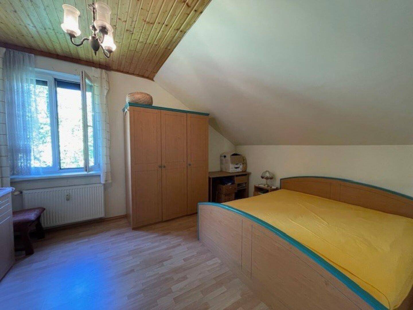 Gemütliches Schlafzimmer im Dachgeschoss mit schräger Holzdecke und großem Fenster.