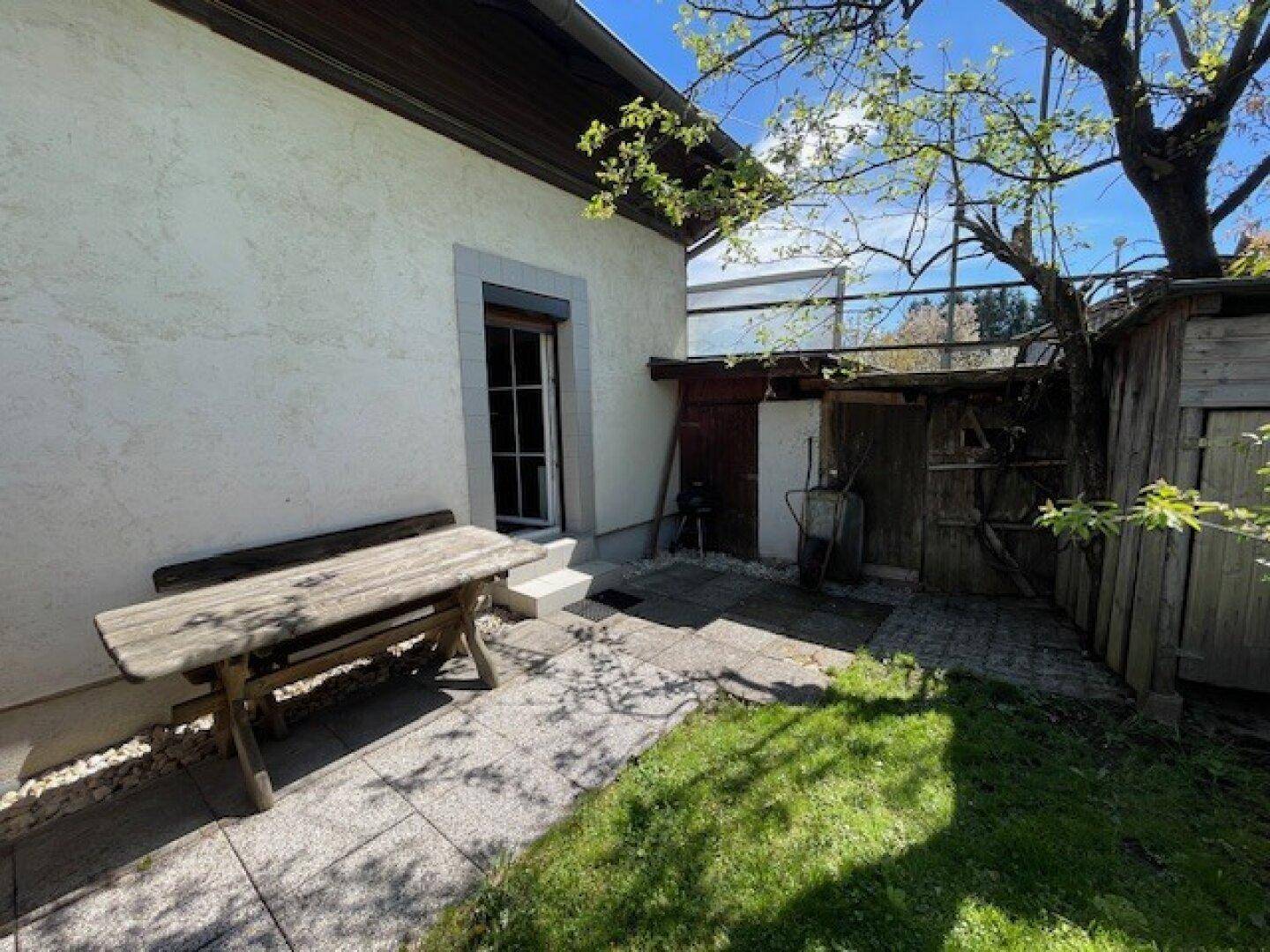 Kleiner gepflasterter Terrassenbereich neben dem Haus mit Holzsitzgelegenheiten und Gartenhaus.