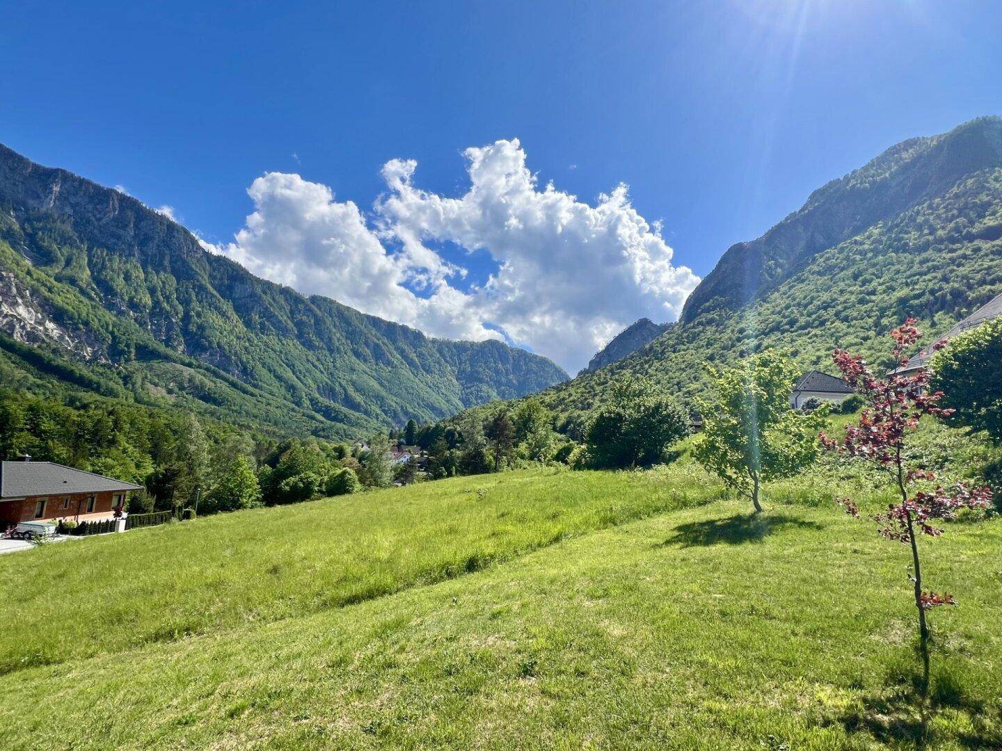 Haus in einer sonnigen Berglandschaft mit grünen Wiesen und Bäumen unter einem weiten Himmel.