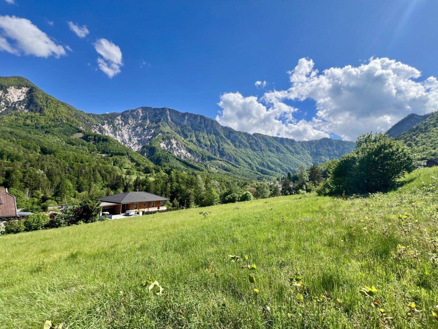 Ein Haus in einer weitläufigen grünen Landschaft mit Bergen und einem bewölkten Himmel im Hintergrund.