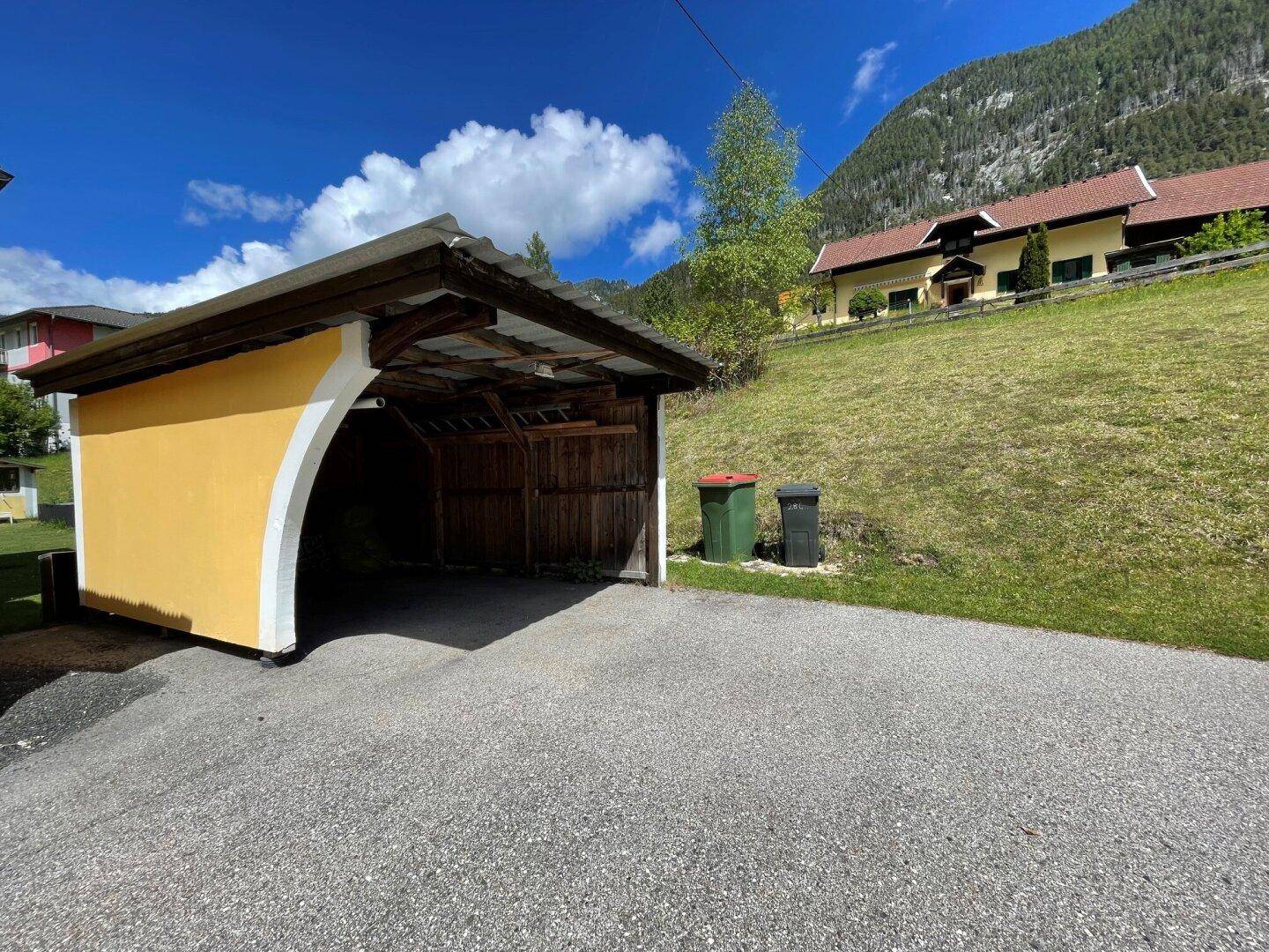 Offener Carport mit Holzdach und asphaltierter Zufahrt.