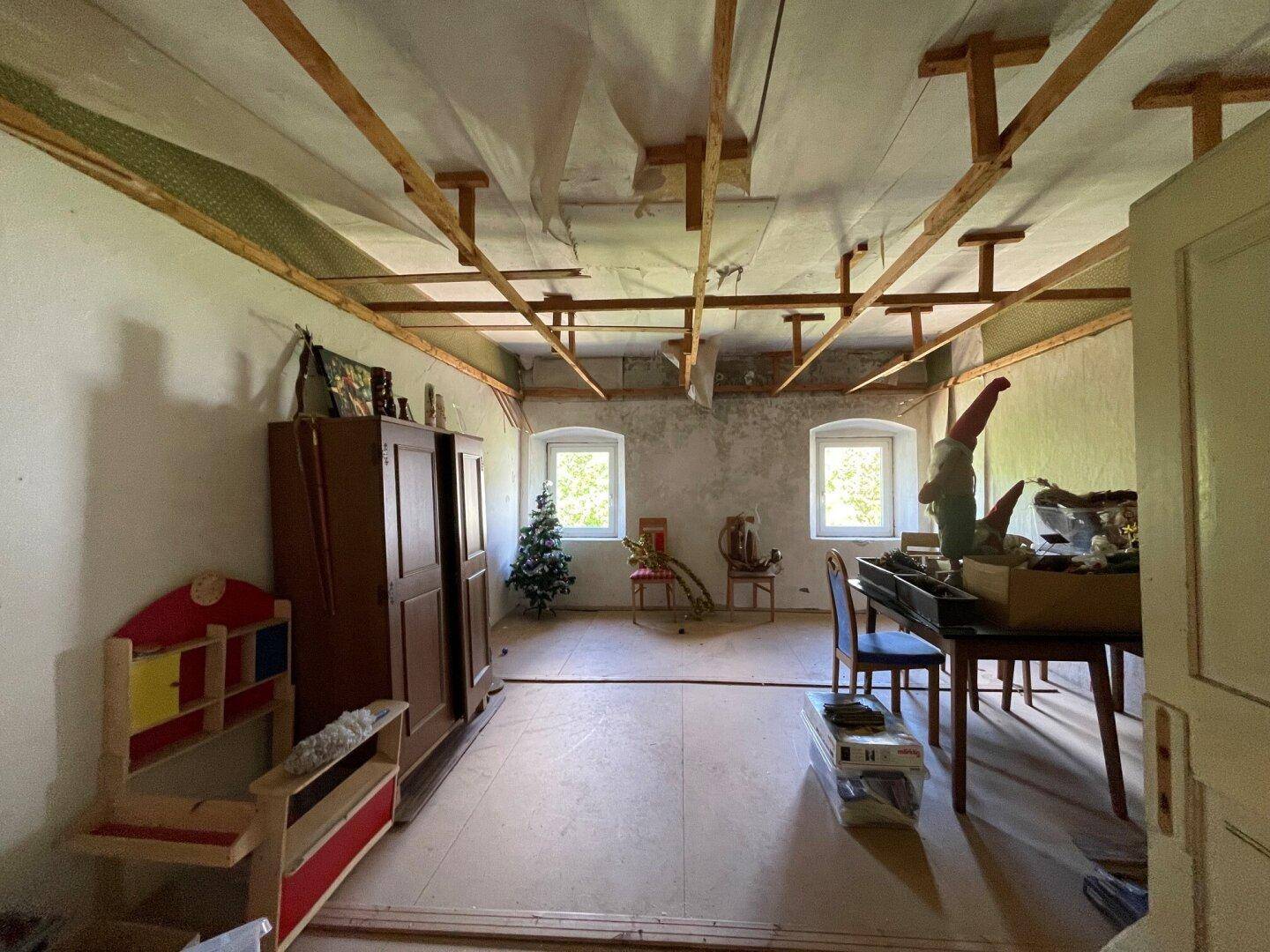 Unfertiger Raum im Dachgeschoss mit Holzbalken und zwei Fenstern.
