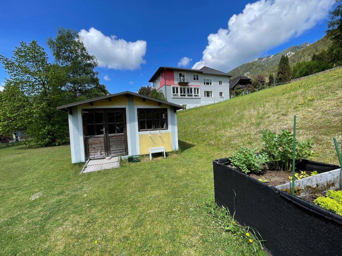 Gartenhaus mit Hochbeet im Vordergrund, Rasenfläche und einem Haus am Hang mit Bergblick.