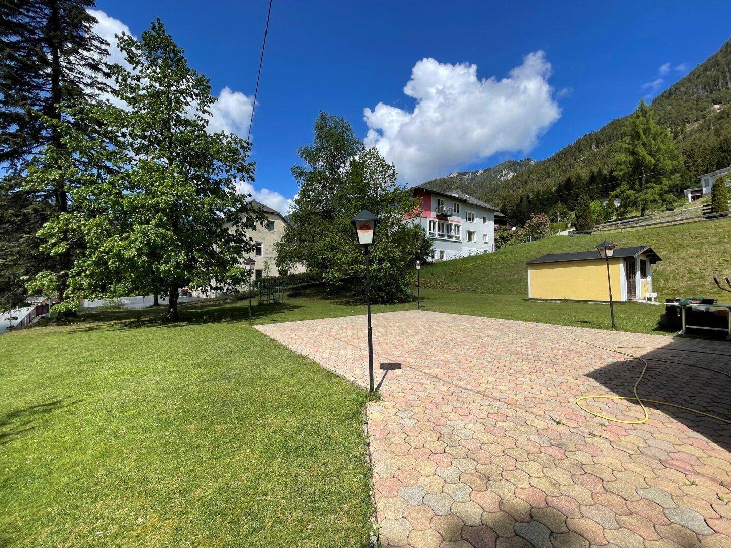 Große gepflasterte Fläche mit Rasen, Bäumen und einem Haus am Hang mit Bergblick.