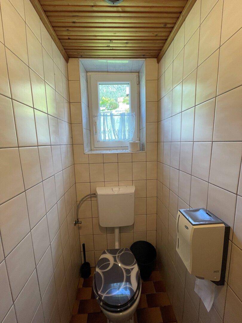 Separate Toilette mit Fliesenwänden, Fenster und gemustertem Toilettensitz.