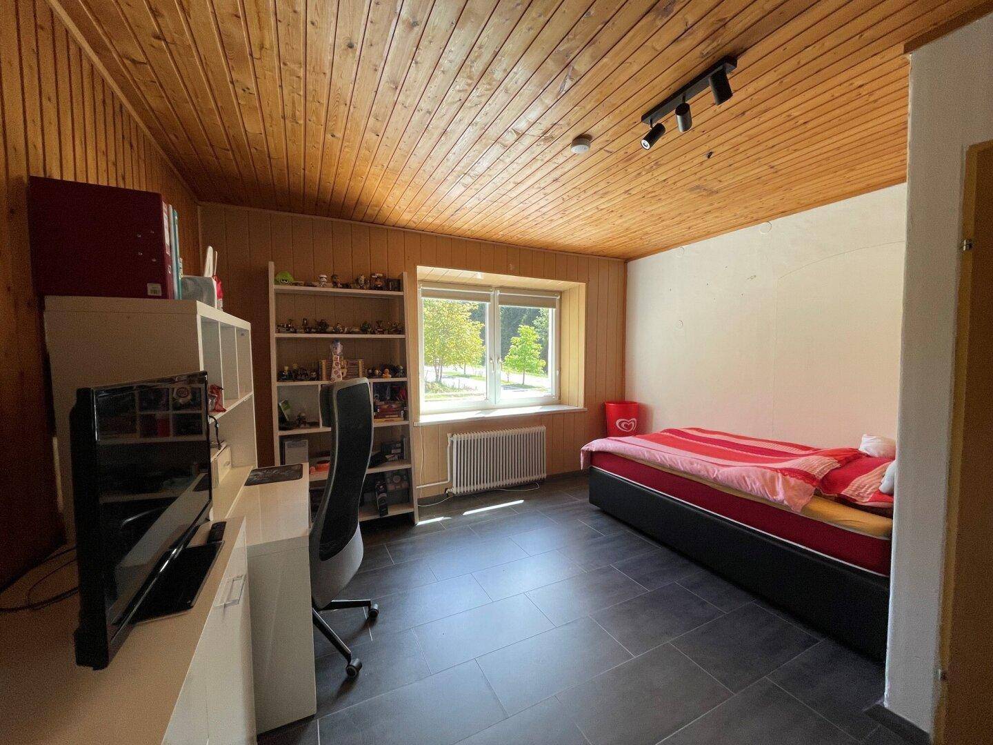 Schlafzimmer mit Holzdecke, dunklem Fliesenboden und Schreibtisch am Fenster.