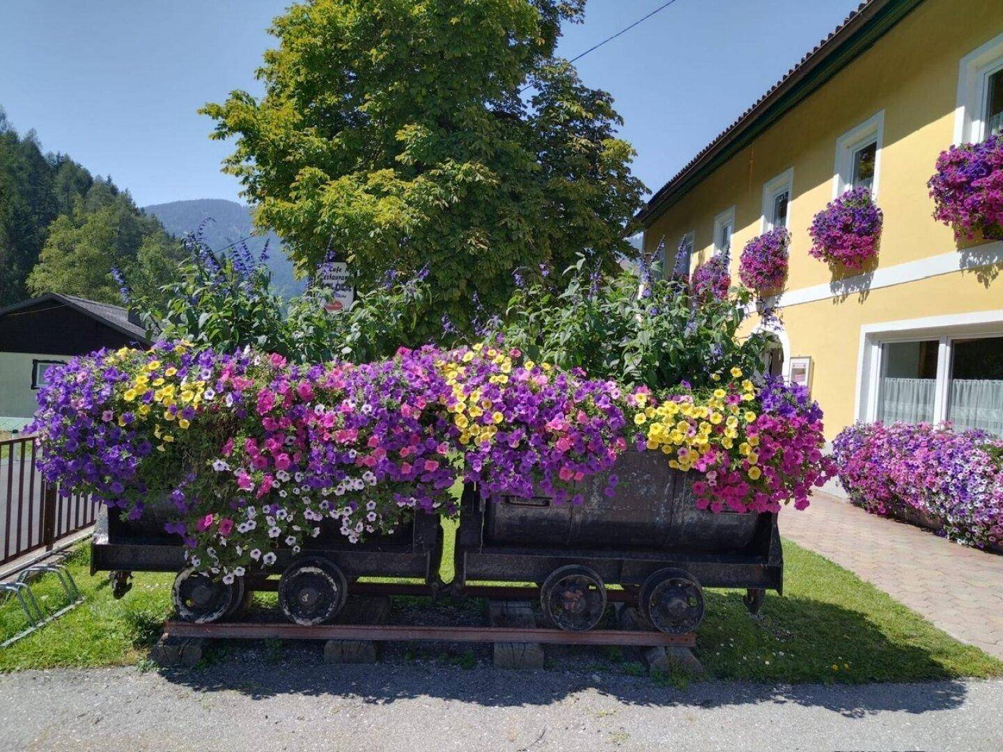 Dekorative Blumenpracht in einem Lorenwagen vor einem gelben Gebäude an einem sonnigen Tag.