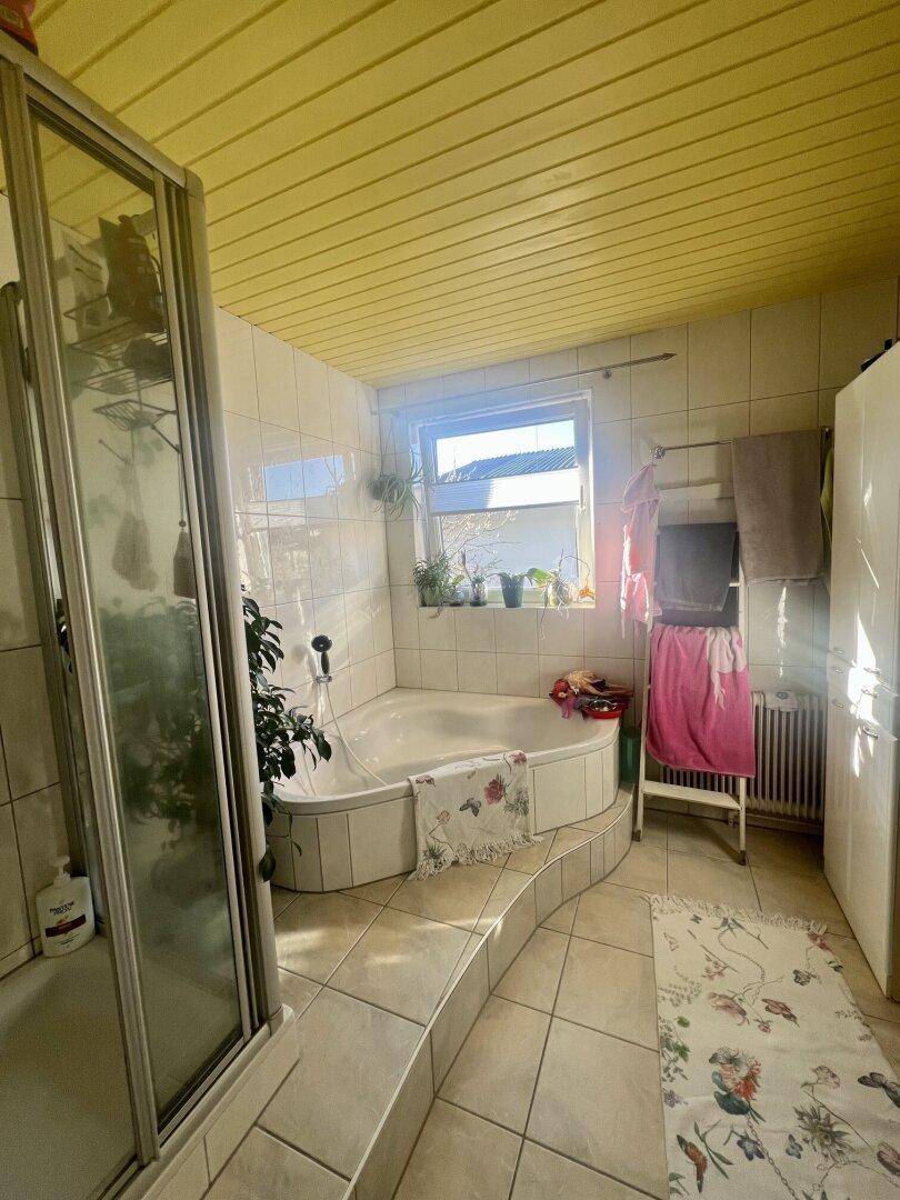 Badezimmer mit Eckbadewanne, separater Duschkabine, Fliesenboden und einem Fenster für Tageslicht.