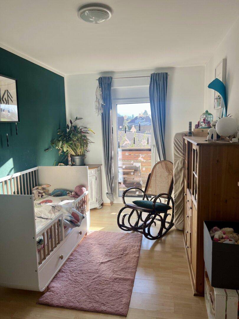 Kinderzimmer mit Babybett, Schaukelstuhl und Zugang zu einem kleinen Balkon mit Blick ins Grüne.