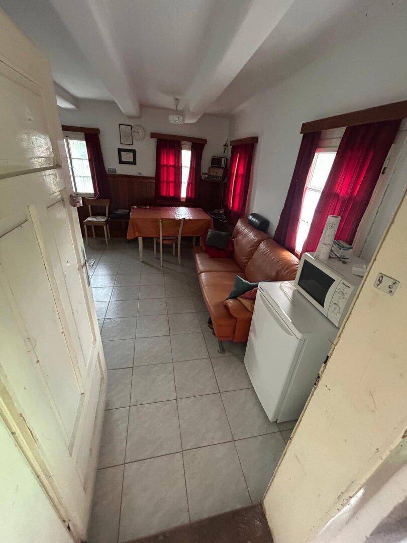 Gemütliches Wohnzimmer mit Fliesenboden, Ledersofa und Esstisch, beleuchtet durch Fenster mit roten Vorhängen.