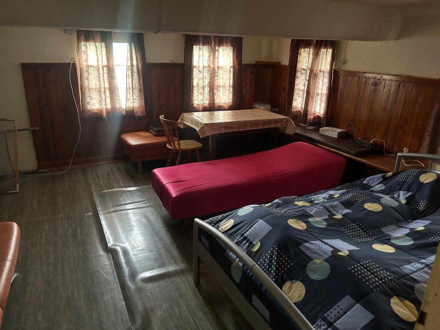 Schlafzimmer mit Holzwänden, zwei Betten und einem Tisch, beleuchtet durch mehrere Fenster mit Vorhängen.