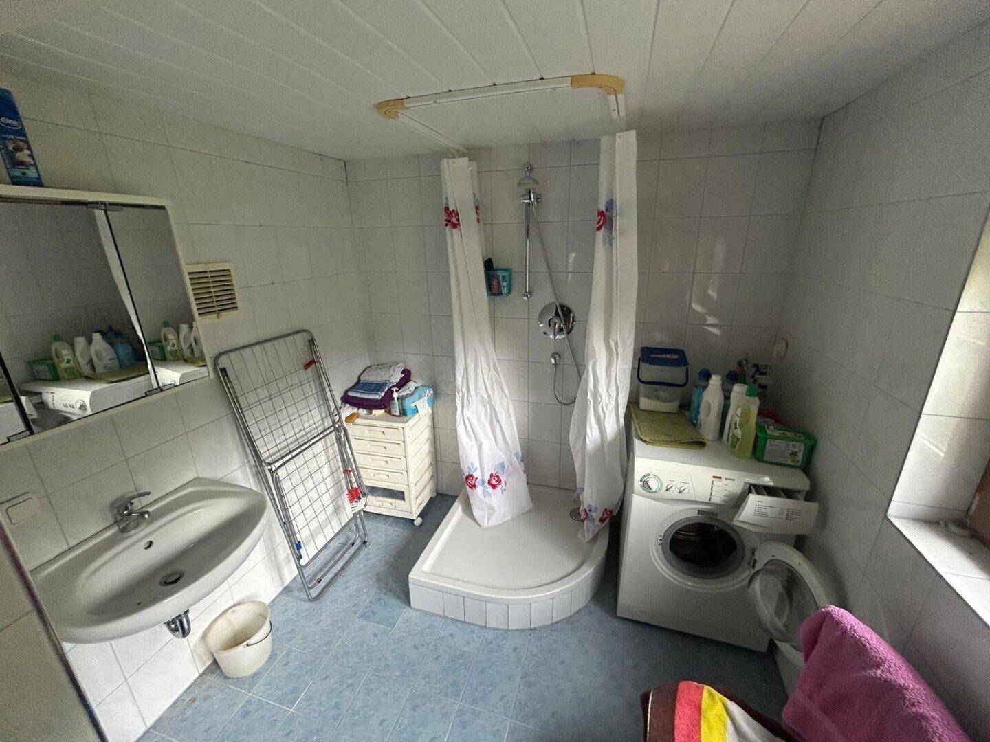 Einfaches Badezimmer mit Dusche, Waschmaschine und Fliesenboden in hellem Ambiente, funktional eingerichtet.
