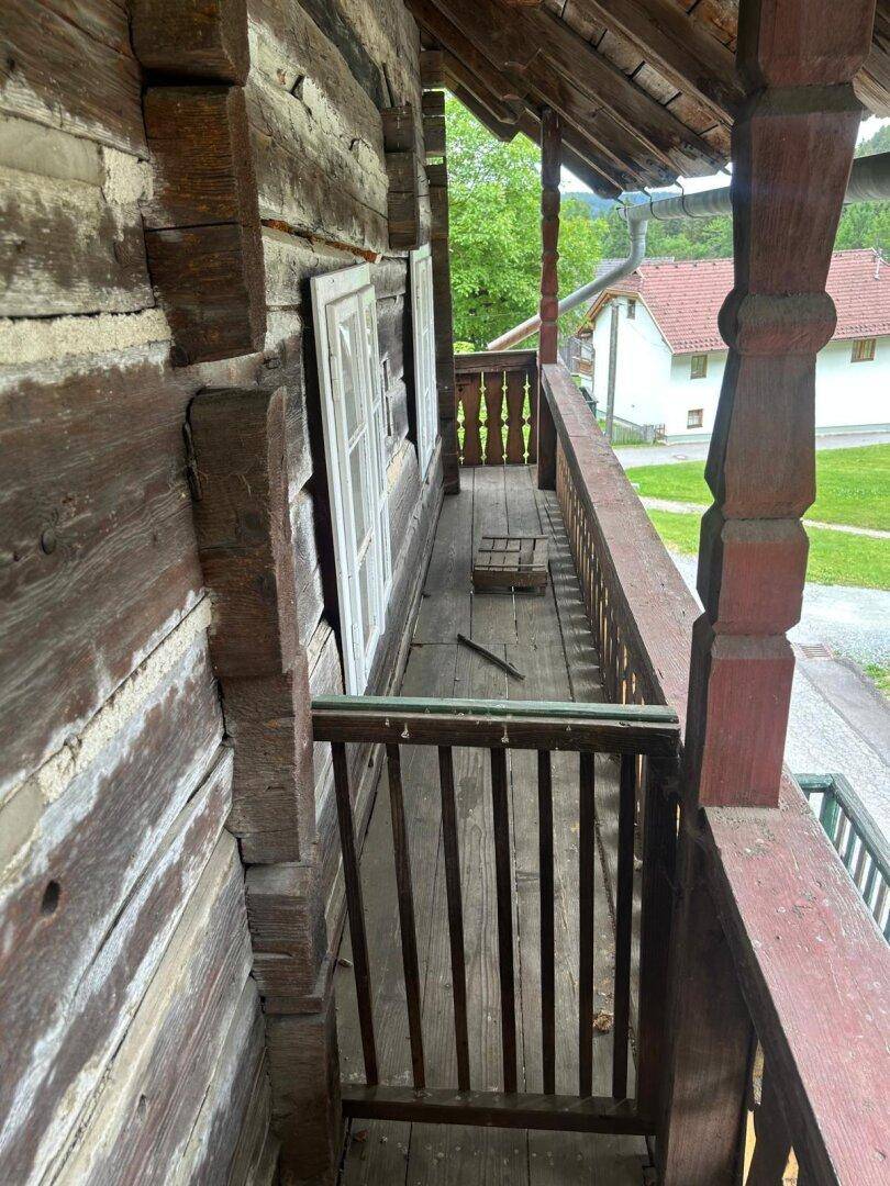 Langer Holzbalkon mit traditionellem Geländer und Blick auf die grüne Umgebung und weitere Häuser.