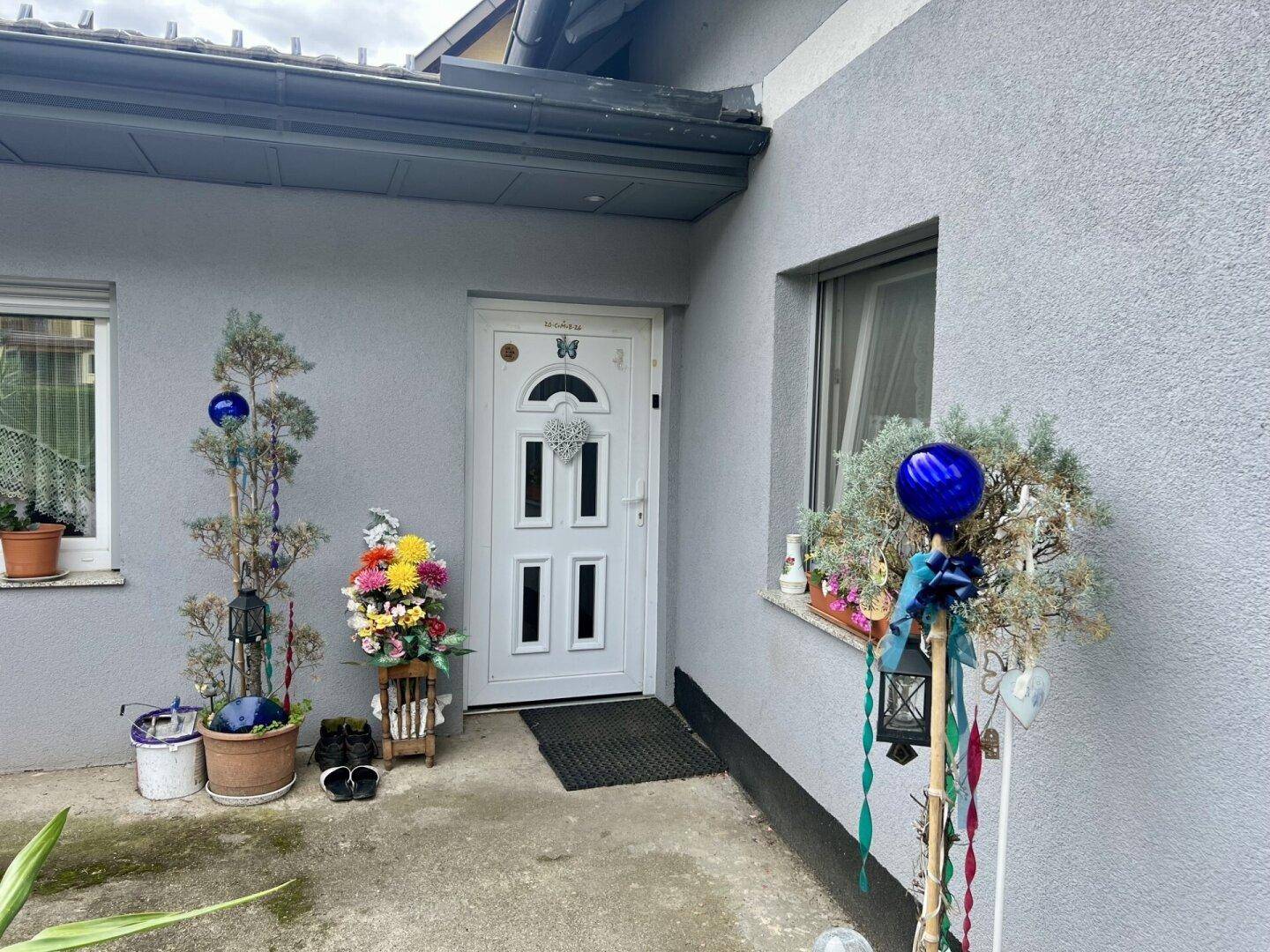 Eingangsbereich des Hauses mit weißer Tür, umgeben von Topfpflanzen und Dekorationen.